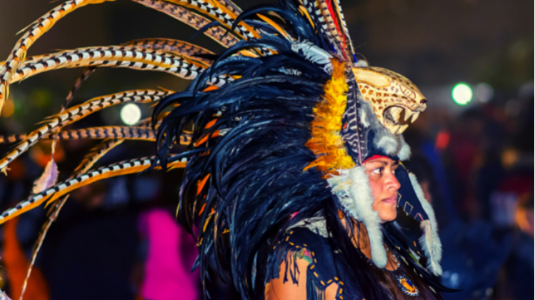 An Aztec Jaguar Warrior running a traditional dance; photo by Jonata Oregón (CC BY-SA 4.0). The photo shows the head and shoulders of an Indigenous person wearing an Aztec headdress with many black feathers emerging from a jaguar head and a dozen longer black and white feathers coming from the center.